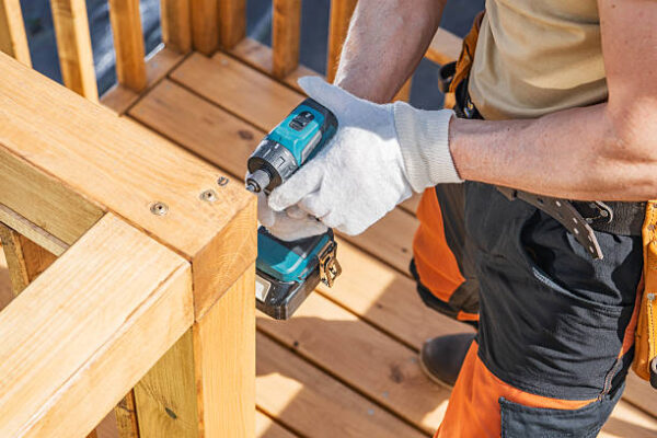 A worker is focused on securing a wooden railing with a cordless drill on a bright, sunny day. Wearing gloves and orange work pants, they are carefully ensuring the railing is stable at a construction site. The warm weather highlights the rich color of the wood and the vibrant surroundings.